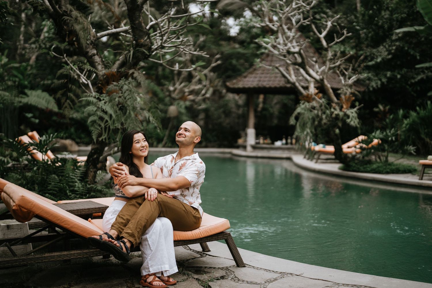 A couple shares a joyful moment while relaxing on a lounge chair by a serene pool surrounded by tropical gardens.