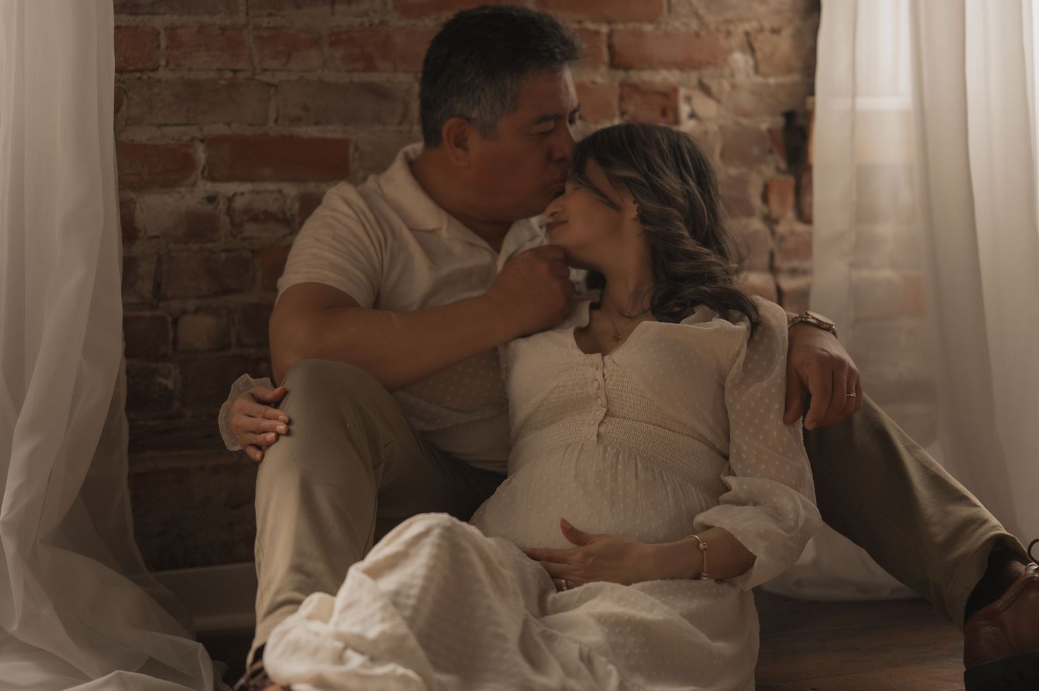 A tender intimate moment between a couple in white clothing against a rustic brick wall with soft natural lighting.