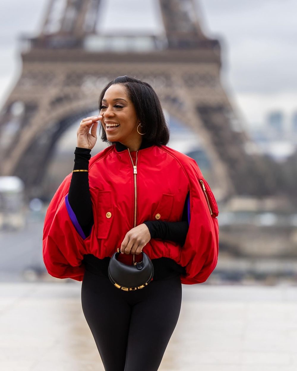 Tourist in vibrant red jacket poses near the iconic Eiffel Tower in Paris on an overcast day.
