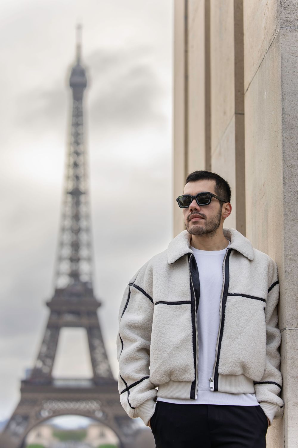 A person wearing sunglasses and light-colored jacket poses against a column with the Eiffel Tower in the background in Paris.