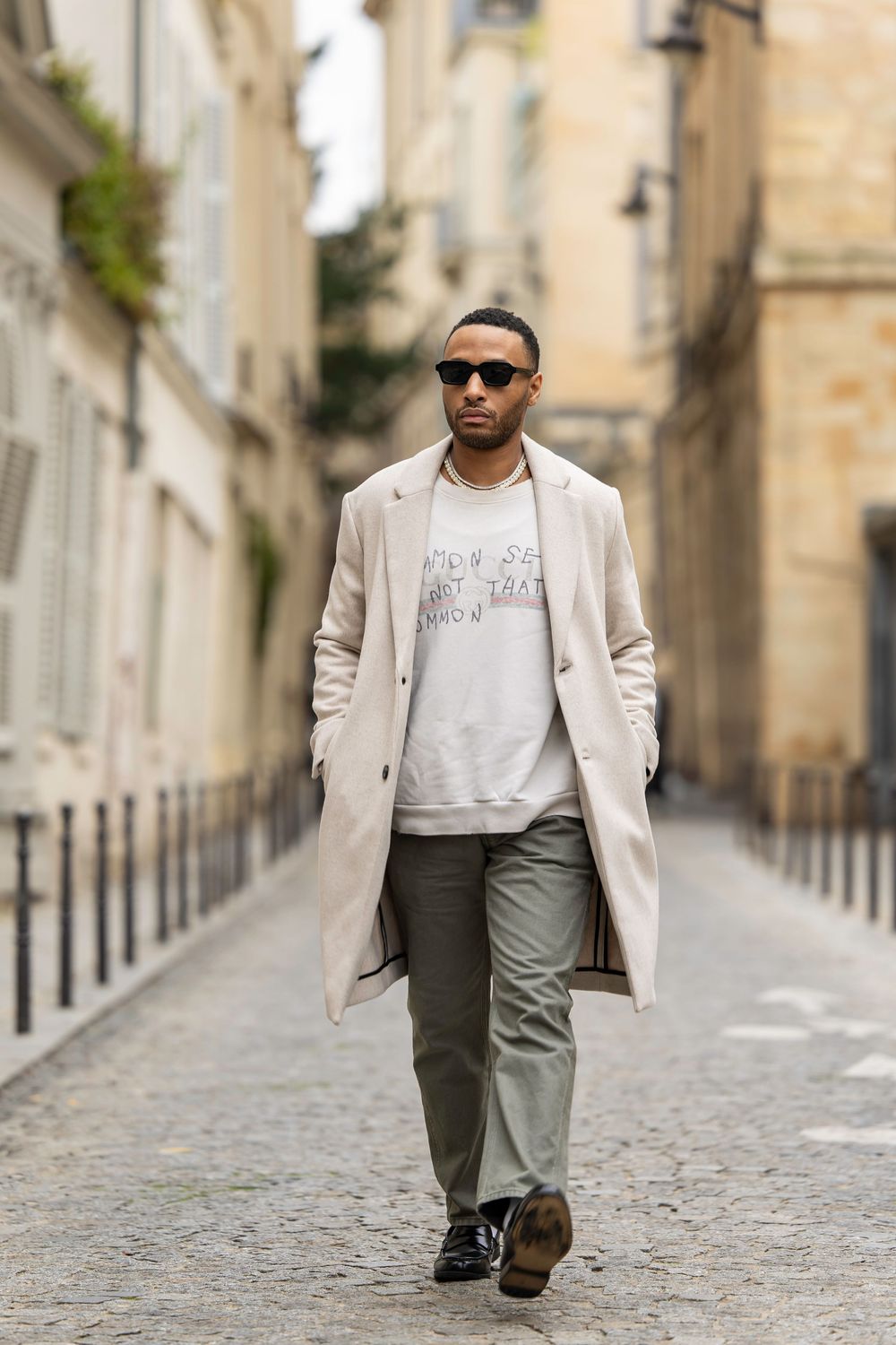 Stylishly dressed person walking on cobblestone street wearing beige coat, green pants and sunglasses in European city.