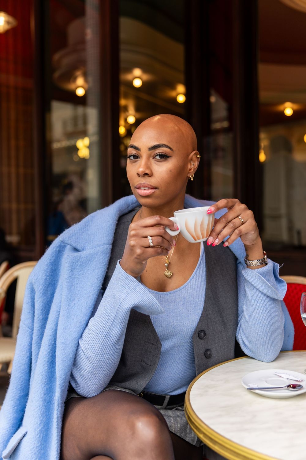 Person in light blue sweater and black blazer sips coffee while sitting at cafe table with geometric backdrop.