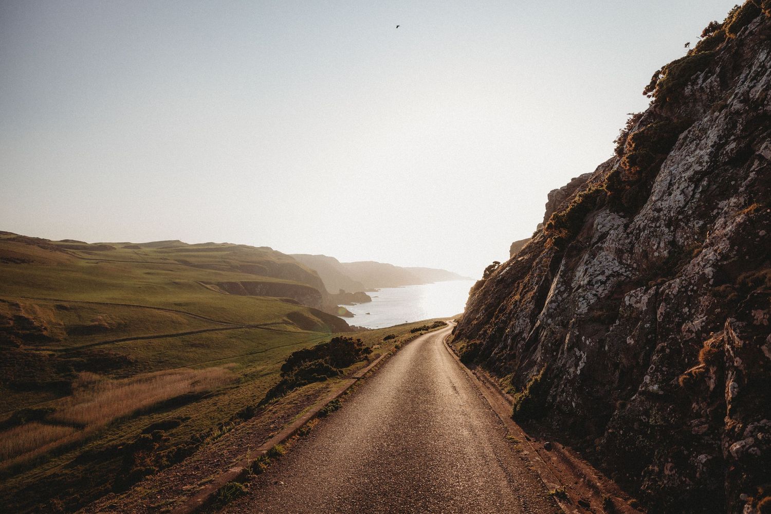 A scenic coastal road winding along rugged cliffs and grassy hills during golden hour with ocean views.