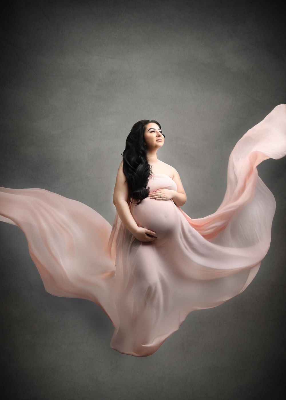 Artistic maternity photo with flowing pink fabric against dark background creating ethereal movement.