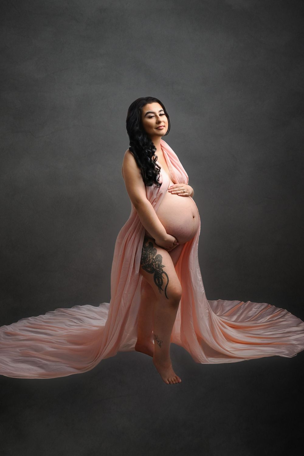 Artistic maternity pose with flowing peach fabric creating graceful lines against grey background.