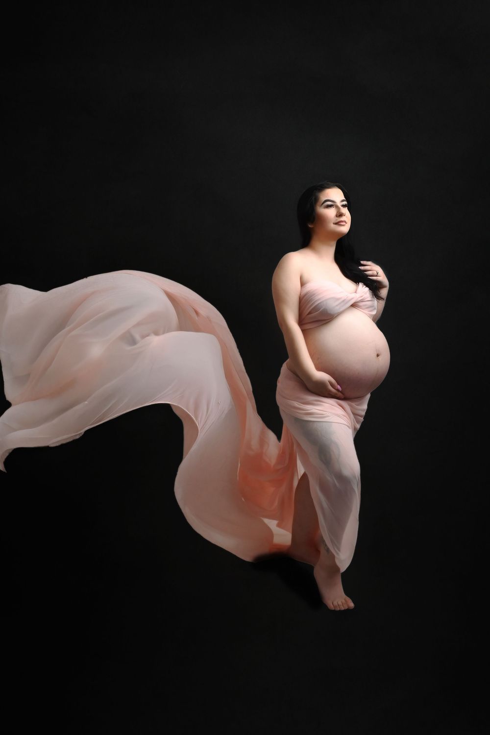 Dramatic maternity silhouette with floating blush fabric against black backdrop.