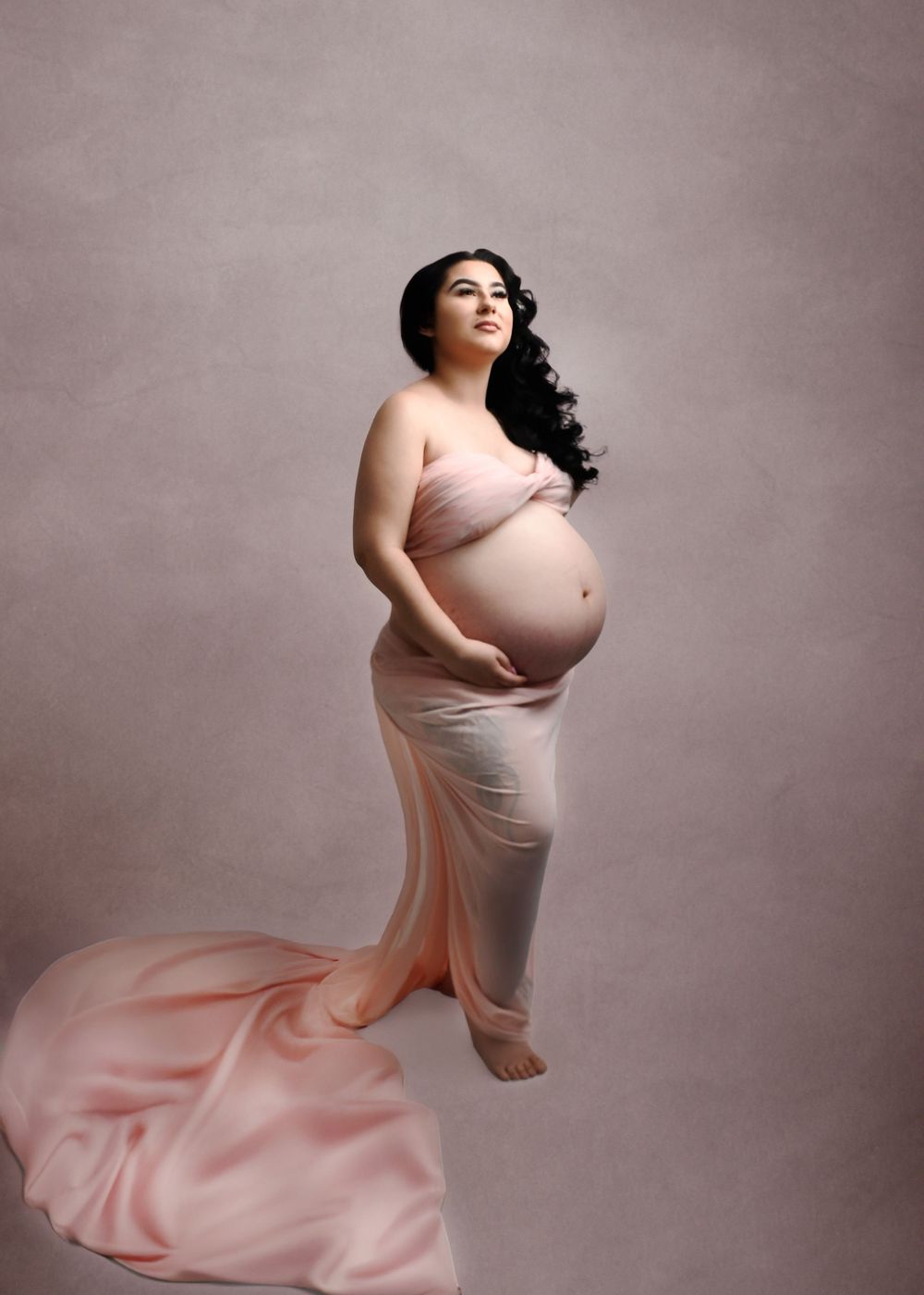 Elegant maternity portrait with long flowing pink dress against neutral backdrop.