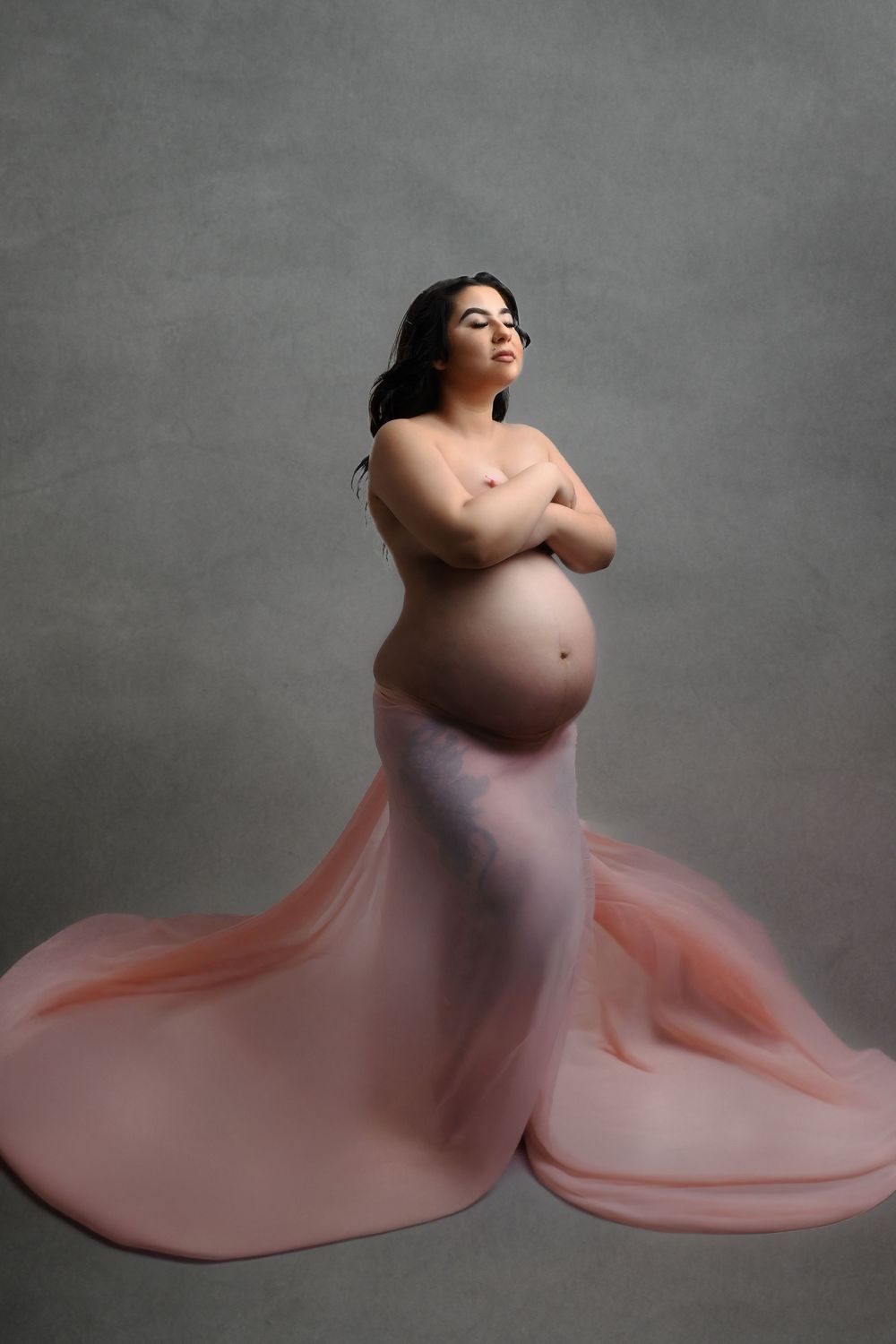 Dramatic maternity silhouette with flowing coral fabric against grey studio background.
