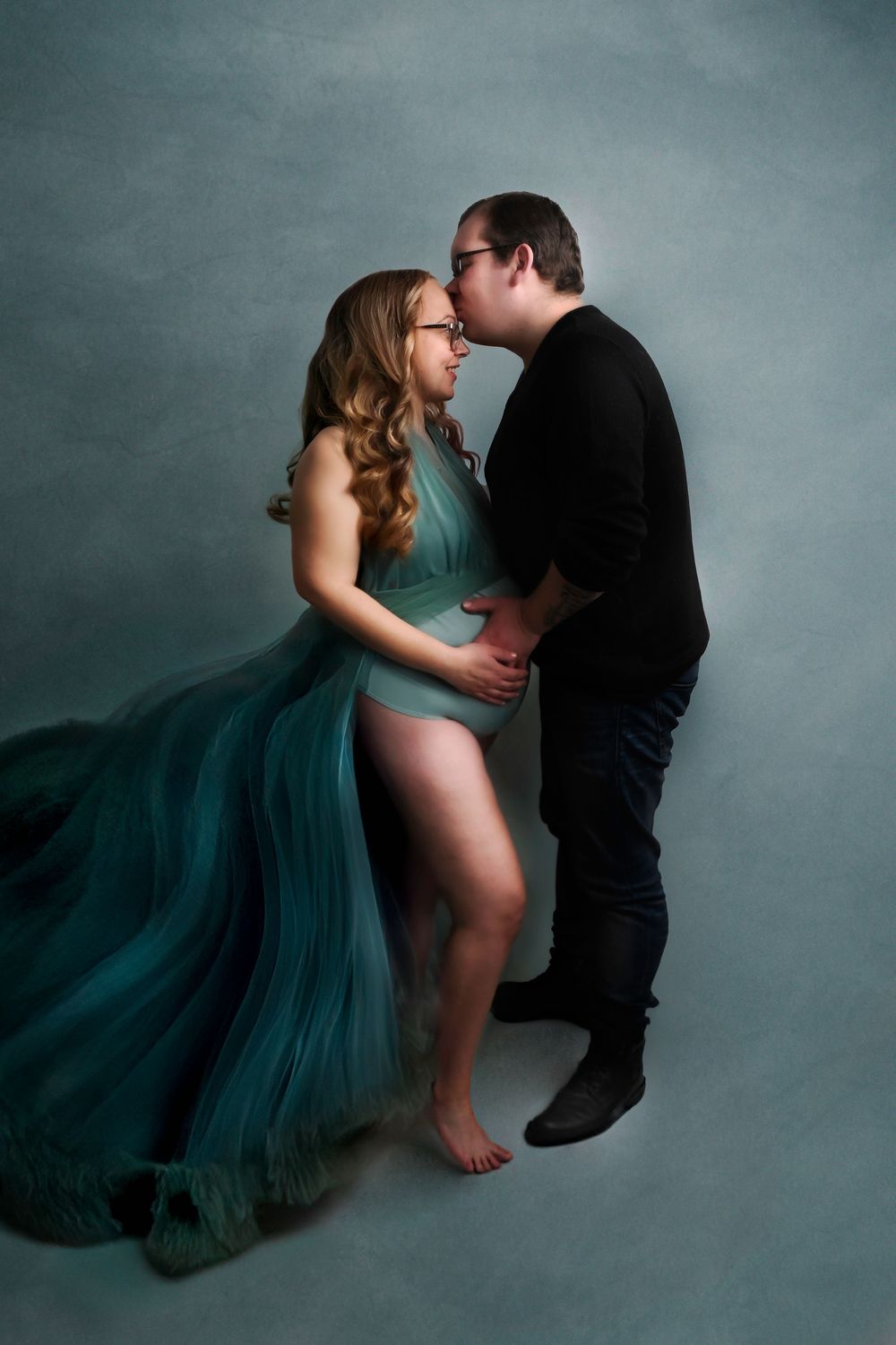 Intimate maternity session with couple in flowing teal gown against grey backdrop.