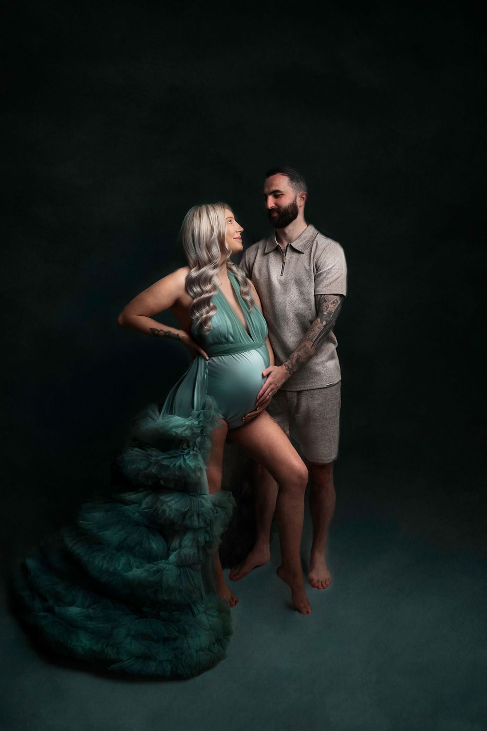 Couple's maternity session with teal flowing fabric creating dramatic effect in dark setting.