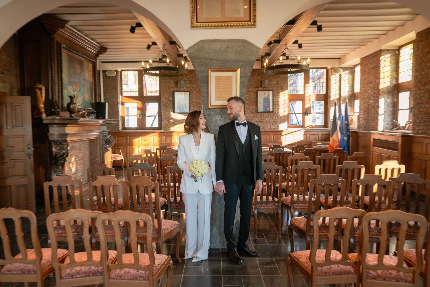 A couple stands in an empty rustic venue with wooden chairs and warm lighting during an intimate wedding ceremony.