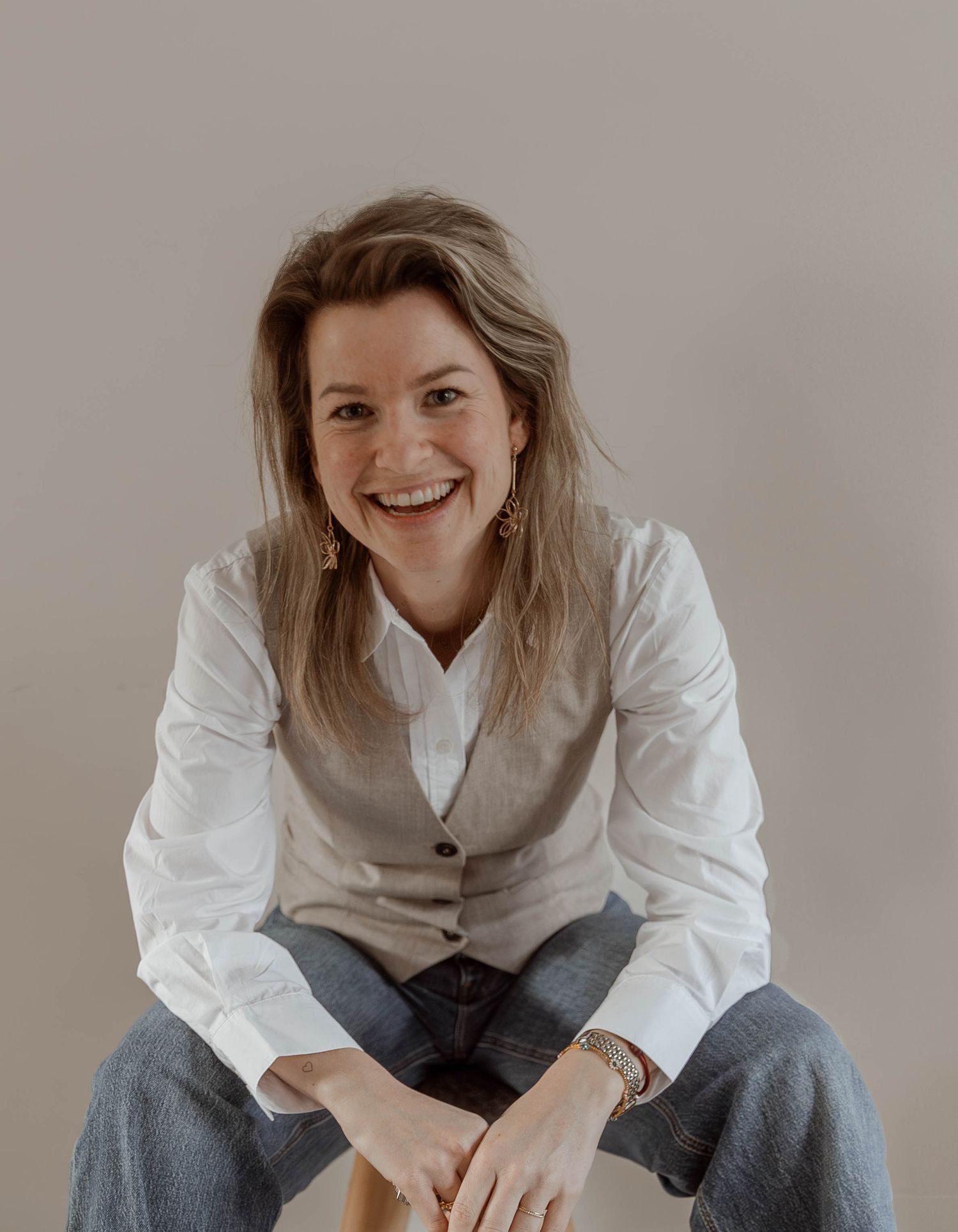 Woman in white button-up shirt and jeans sits casually while smiling in a minimalist setting.