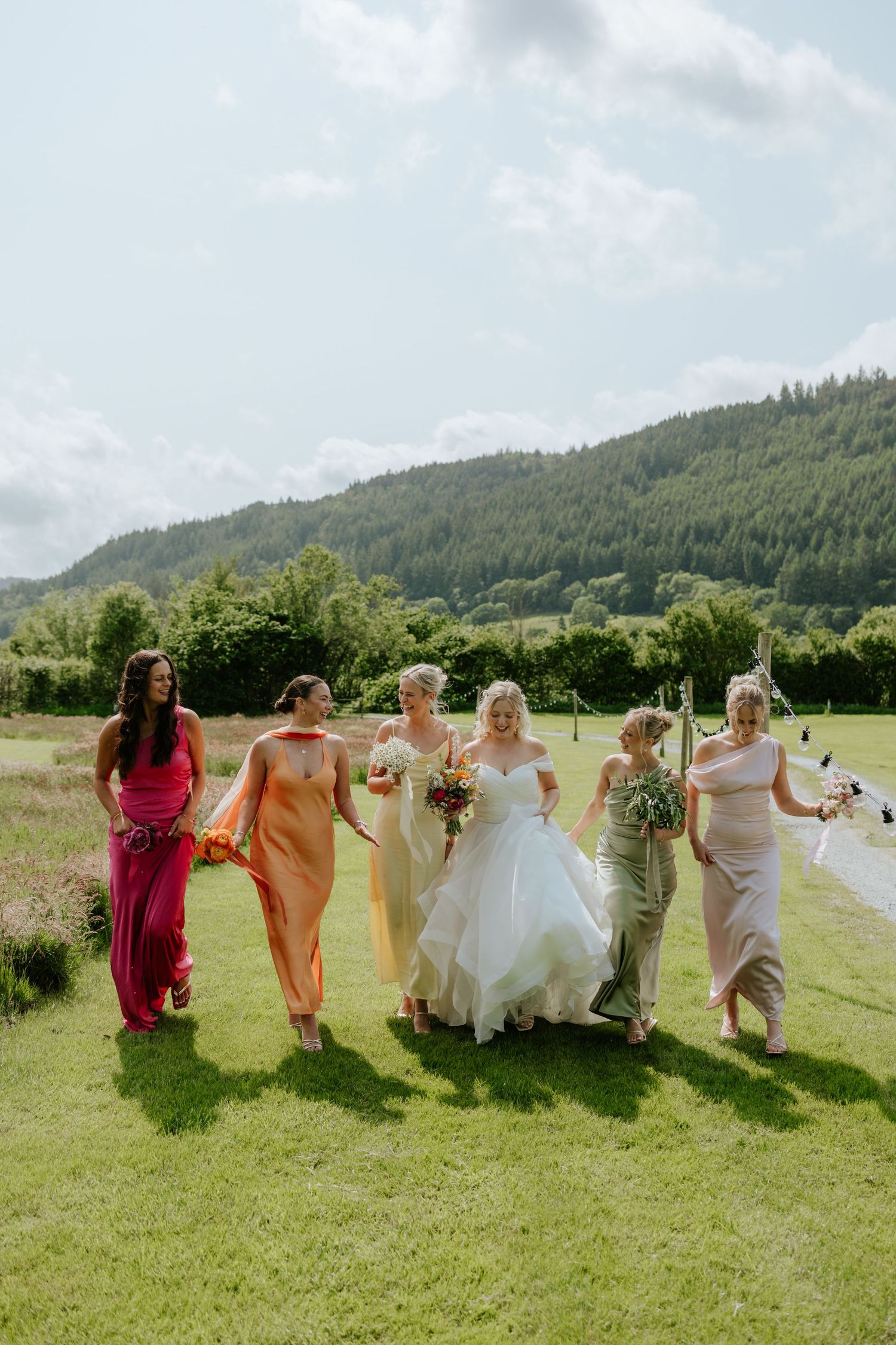 A bridal party in colorful dresses stands together on a grassy lawn with mountains in the background, holding bouquets.
