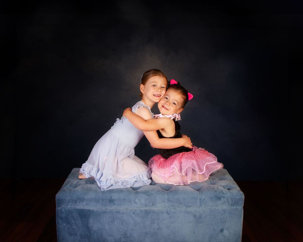 Two children in tulle dresses share a heartwarming embrace while sitting on a blue ottoman against a dark backdrop.