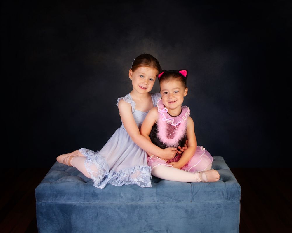 Two young friends in pastel dresses pose together on a blue ottoman against a dark studio background.