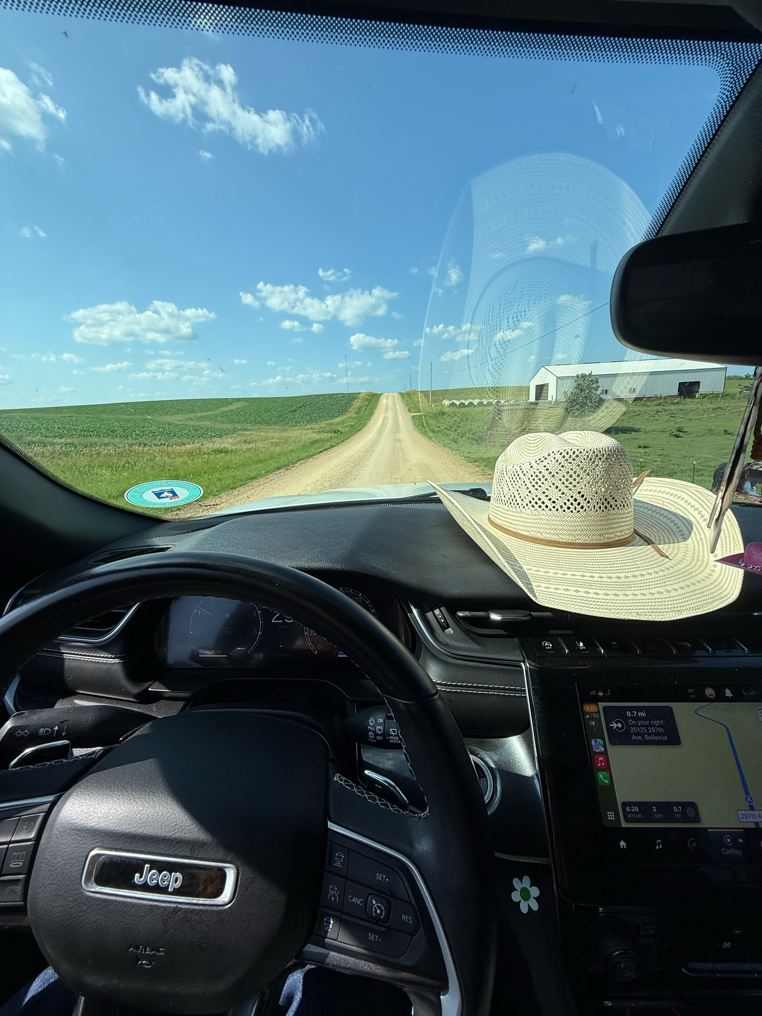 View through car windshield of dirt road stretching across green prairie with blue sky and straw hat on dashboard.