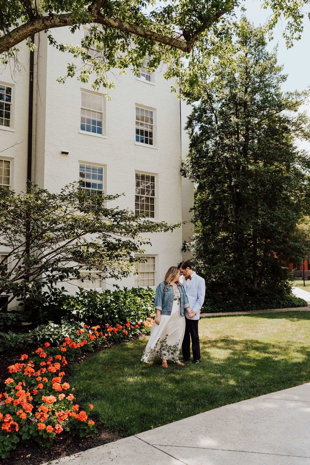 A couple shares a romantic moment on a lawn with orange flowers and a white building in the background at the Gettysburg College Campus.