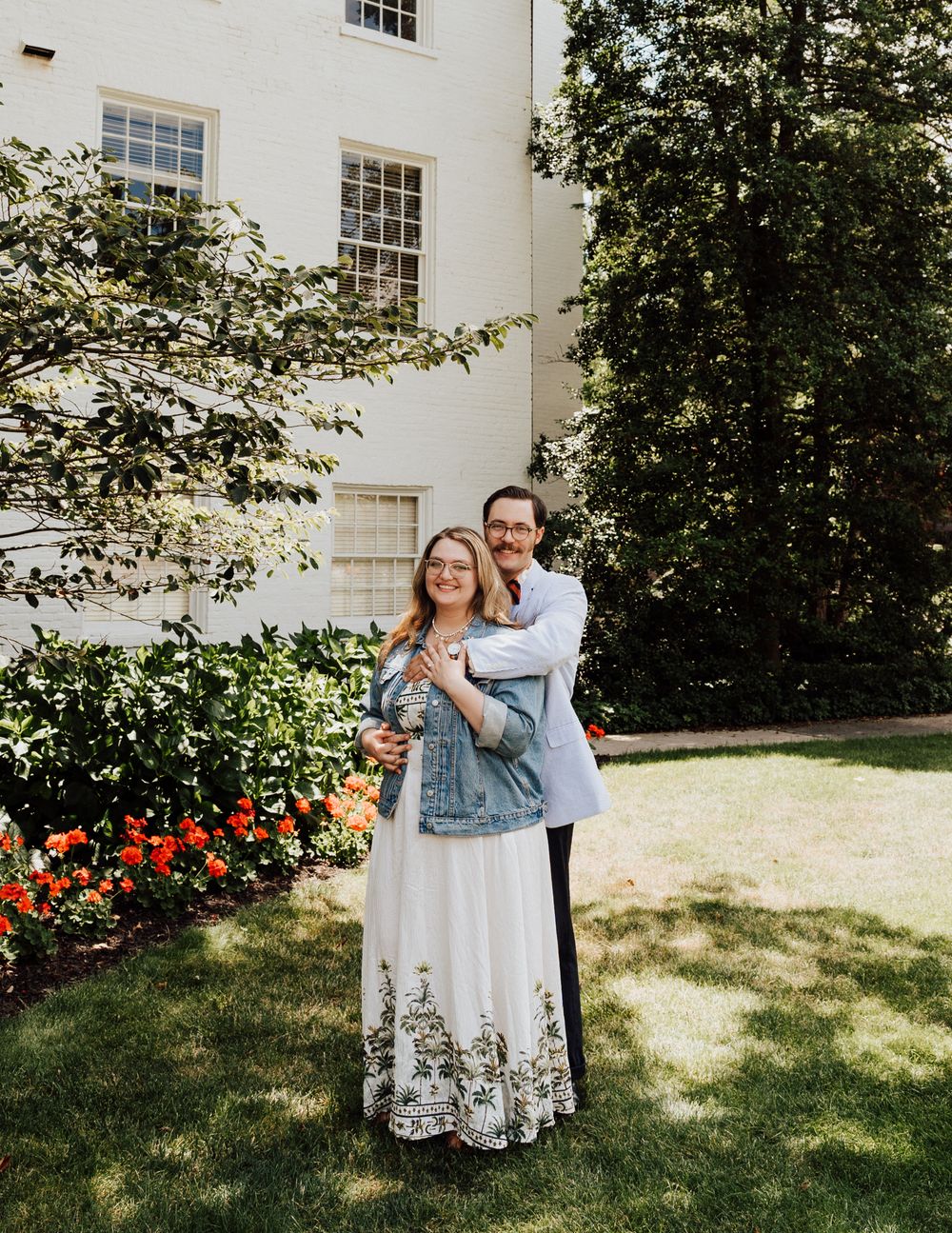 A couple embraces in front of a white colonial style building with red flowers in the garden at Gettysburg College Campus.