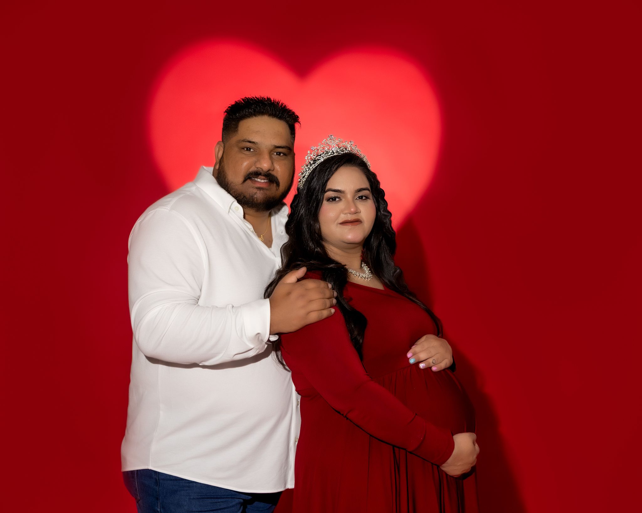 A couple poses together against a red backdrop with a heart shape, wearing white and red outfits for a romantic photo.
