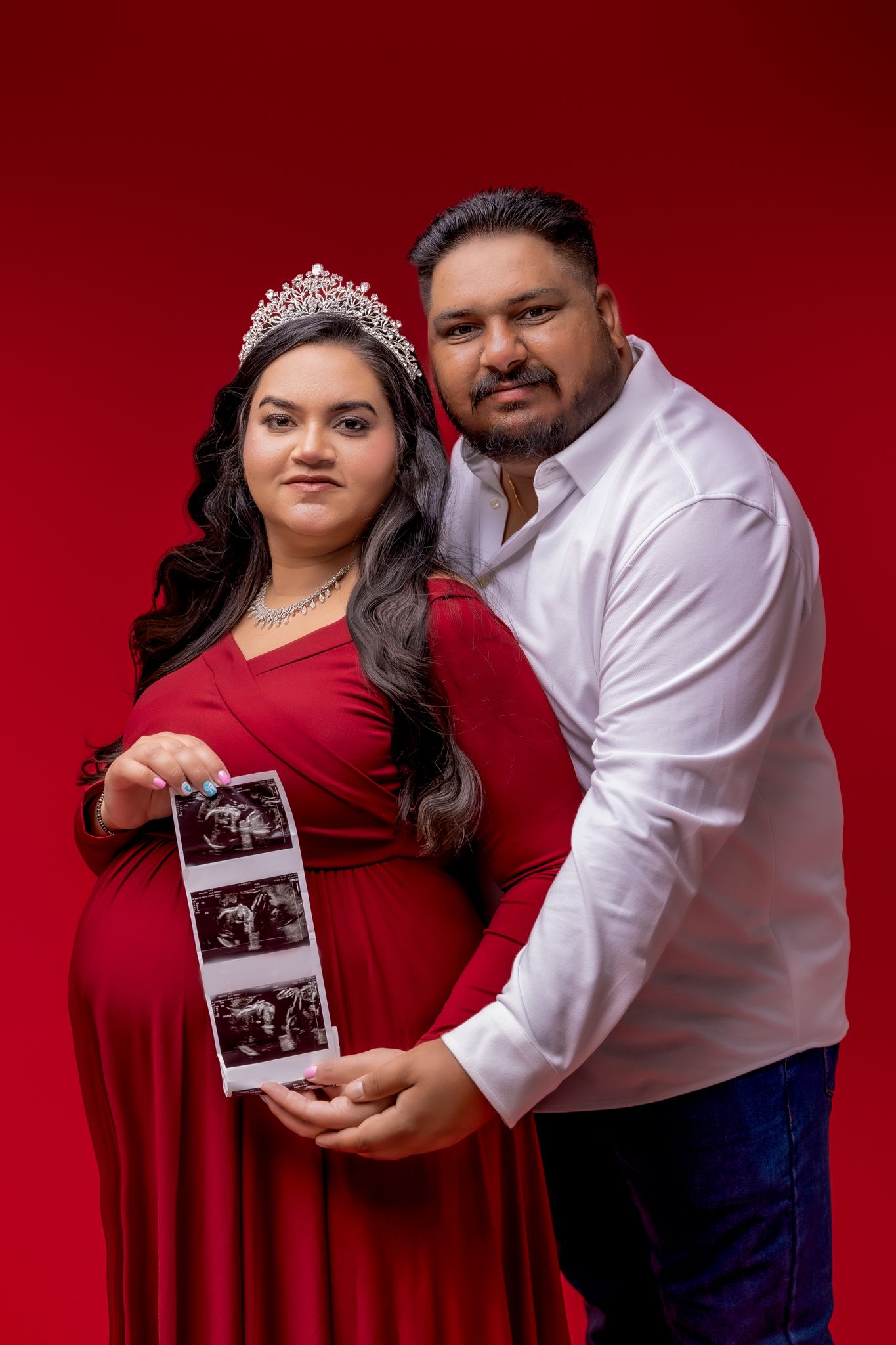 Pregnancy announcement portrait with couple in red and white outfits holding ultrasound photos against crimson backdrop.