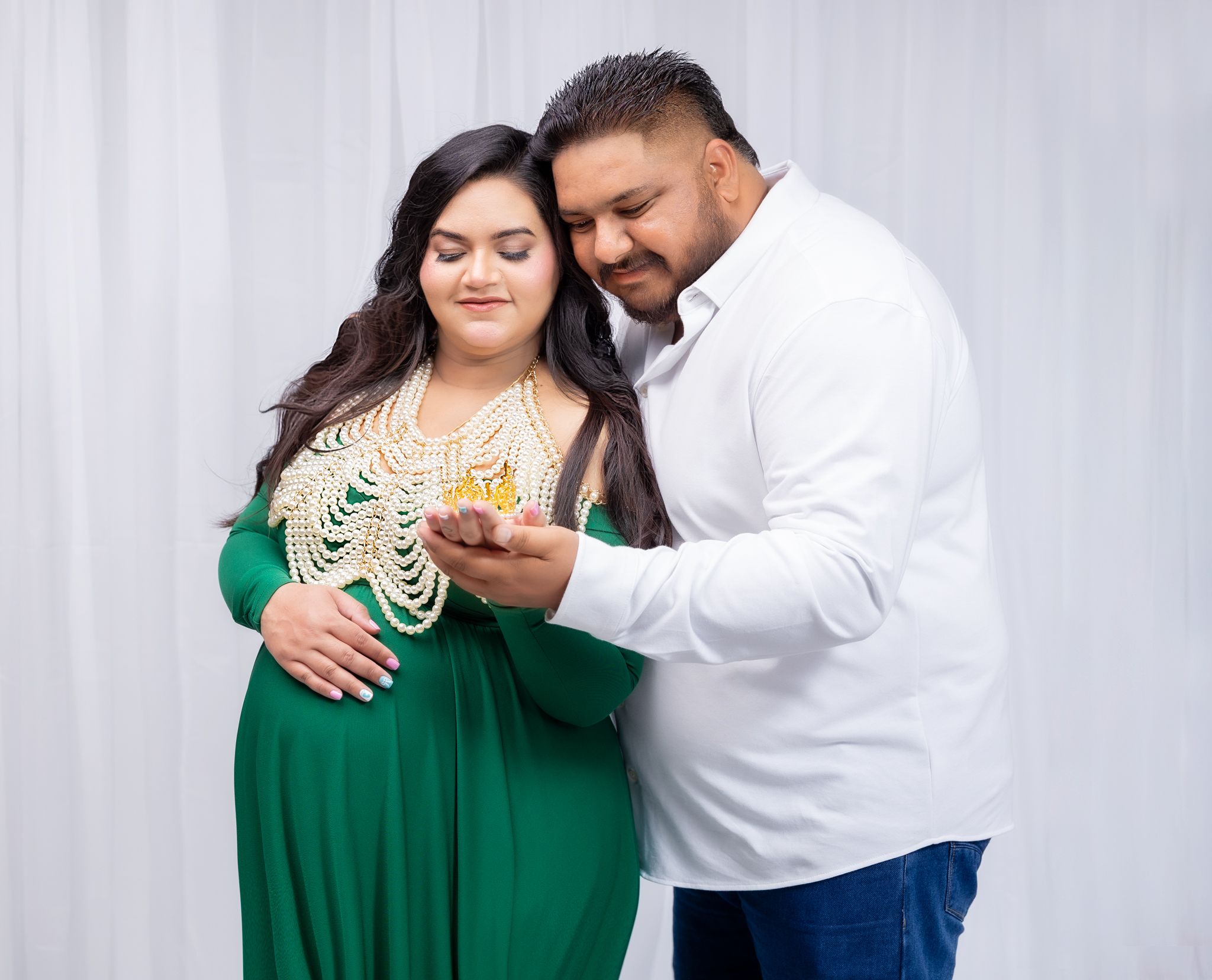 Couple in green dress and white shirt embracing in studio photo session against light gray backdrop.
