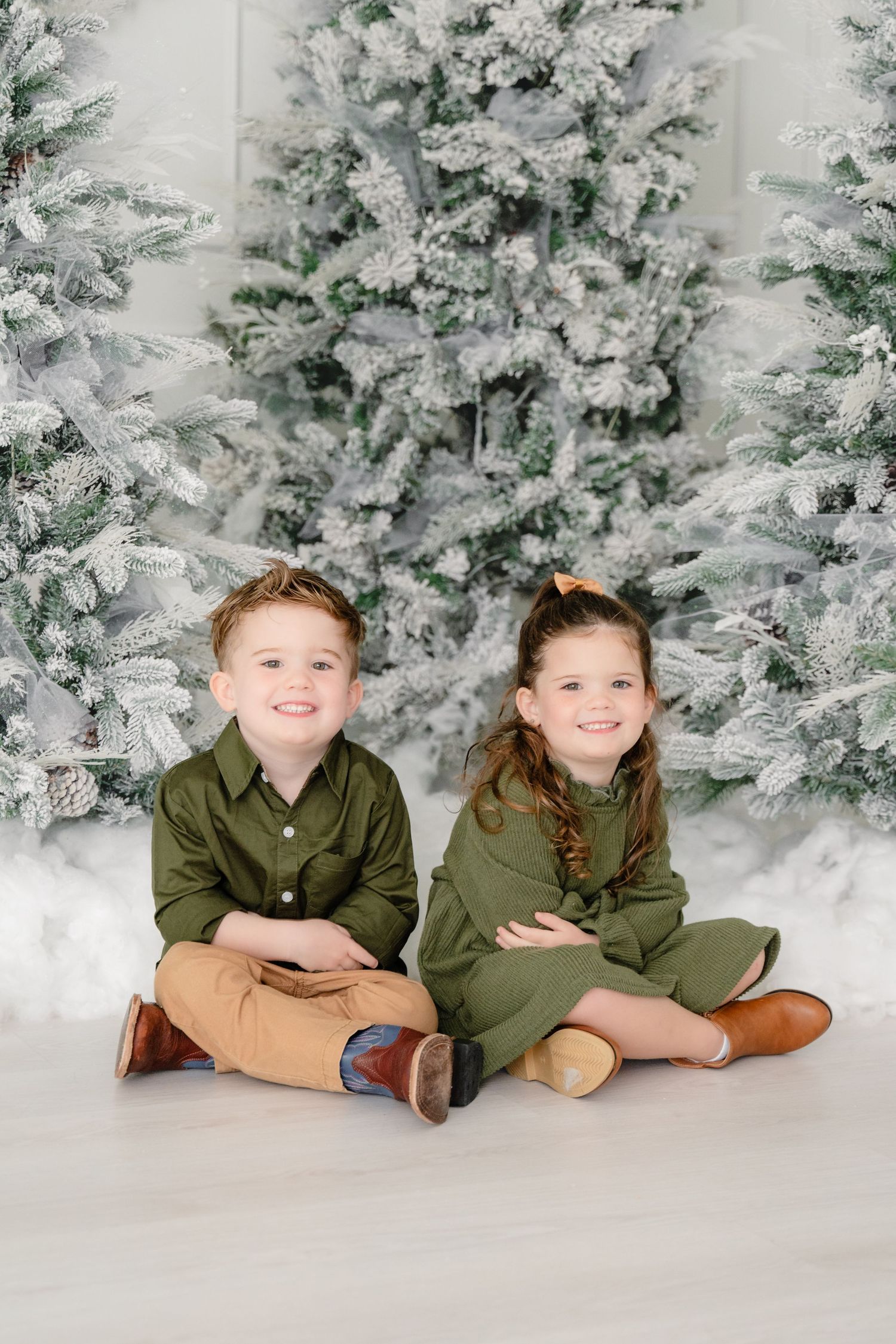Two children sitting together in matching dark green outfits against a snowy winter wonderland backdrop.