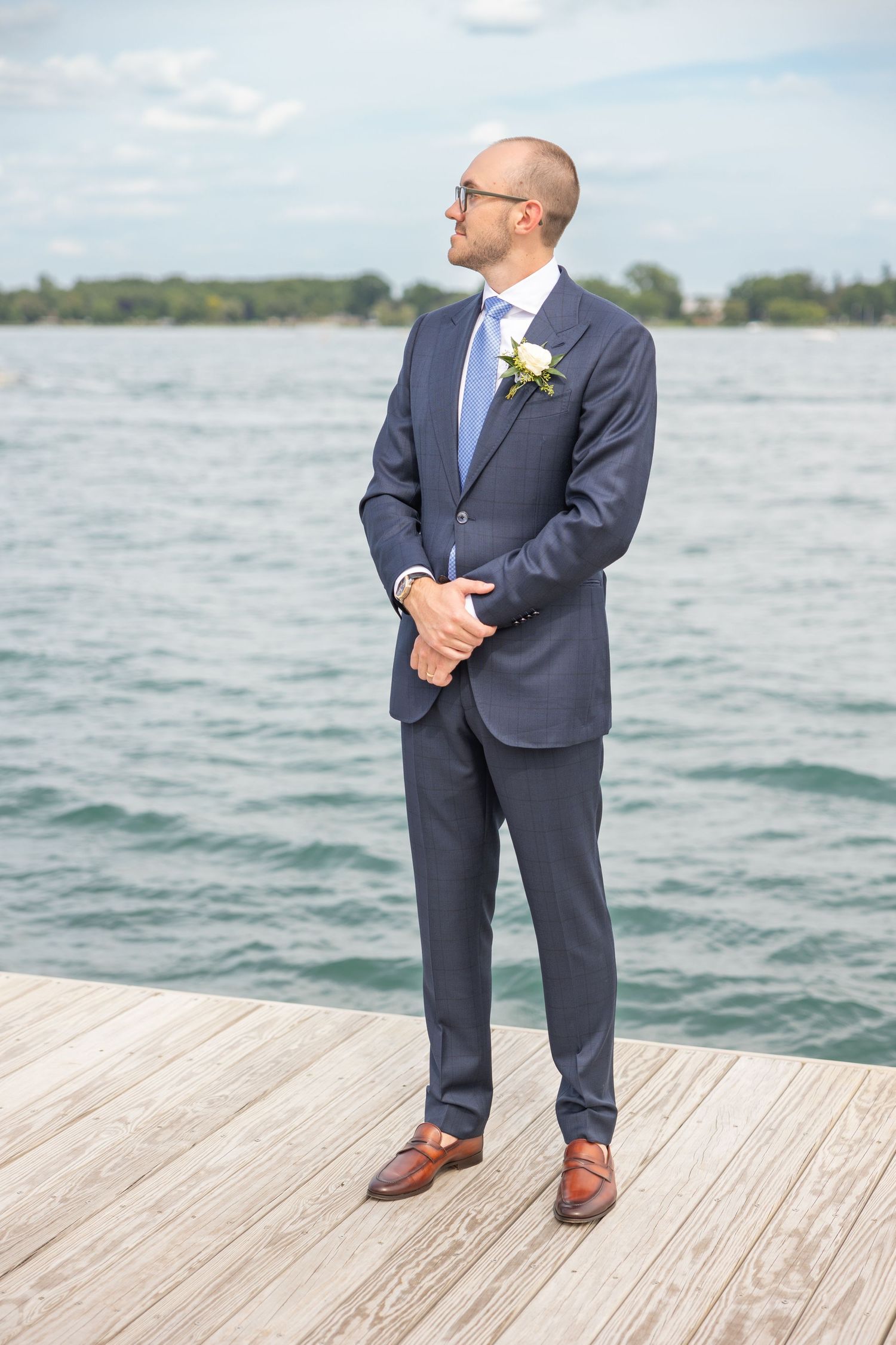 Someone in a navy suit with a boutonniere stands on a wooden dock by a lake on their wedding day. Rachel Martinez Photography Novi MI