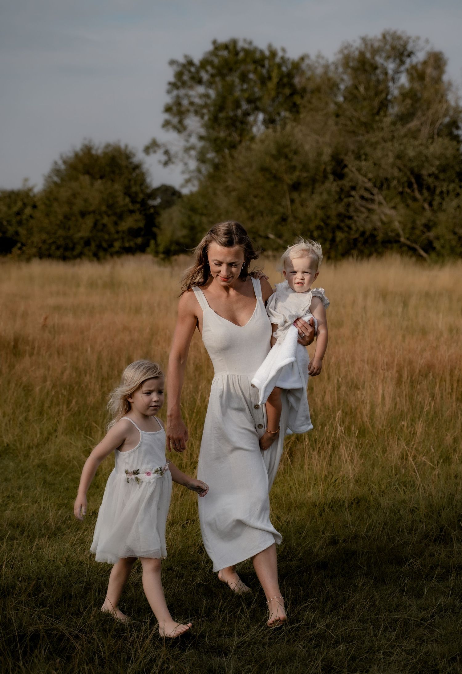 Mother walking barefoot through a meadow with her two young daughters during an outdoor lifestyle family session in Derbyshire, embracing sl