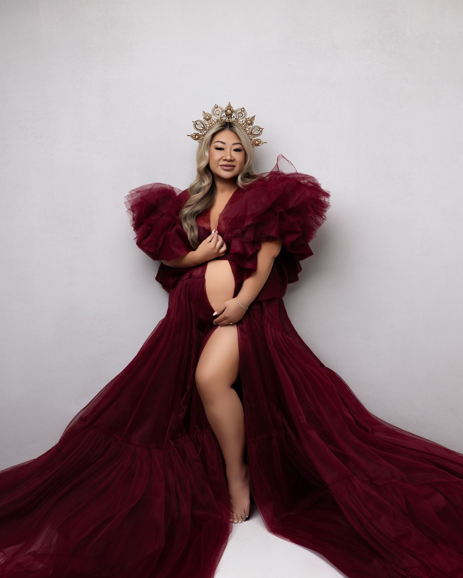 A figure poses gracefully in a flowing burgundy tulle dress against a light gray studio backdrop.