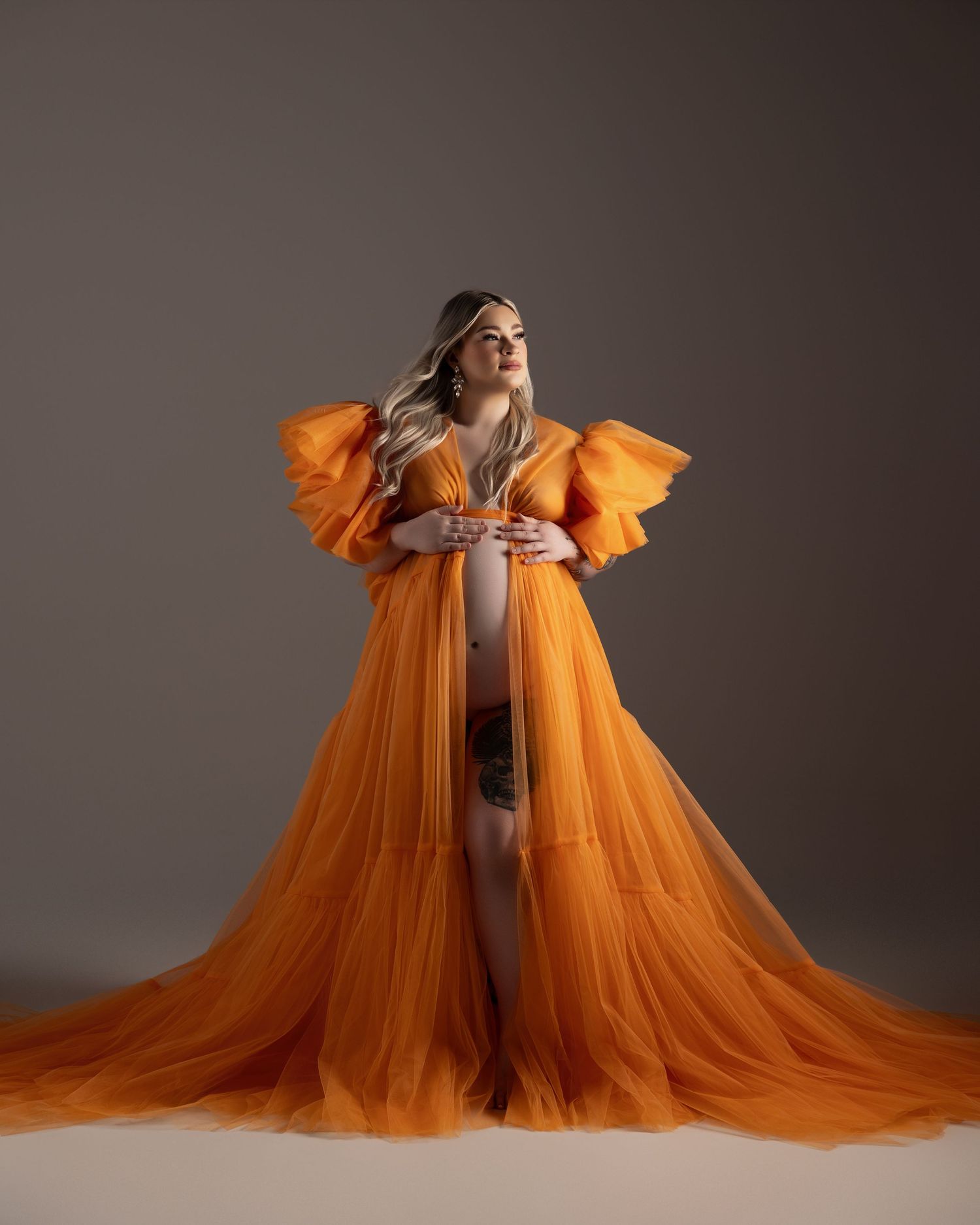 Ethereal orange tulle maternity gown with dramatic flared sleeves and flowing train on gray studio backdrop.