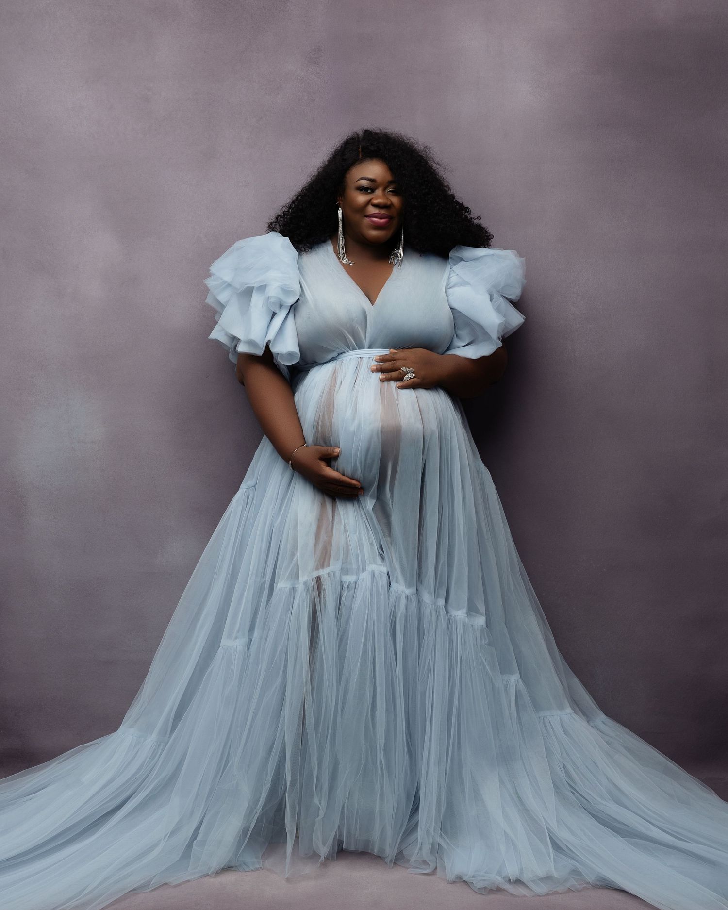 Fashion portrait of a model in a flowing powder blue tulle gown sitting against a gray studio backdrop.