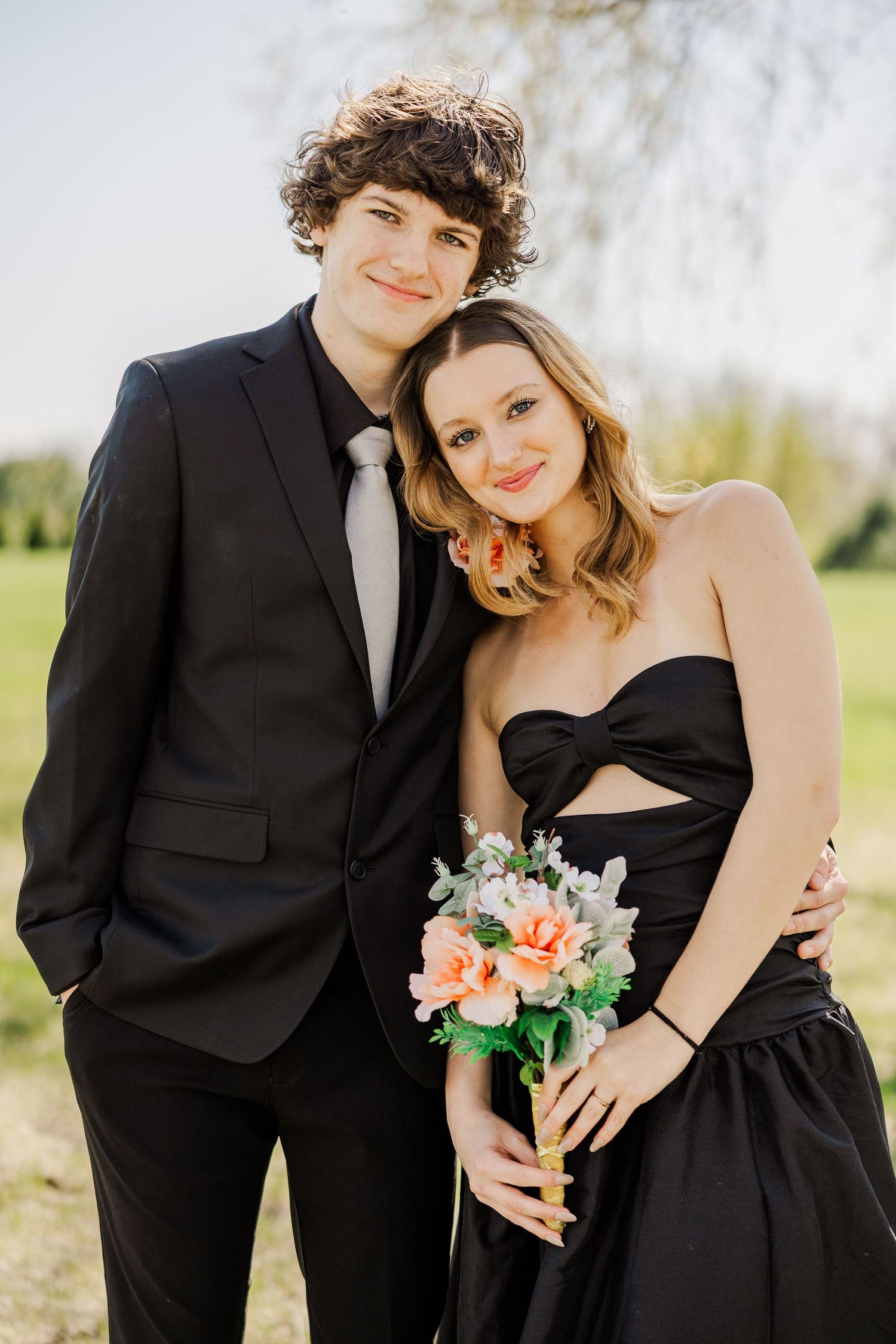 A couple in formal attire poses outdoors, with a black dress and suit, holding a colorful bouquet of flowers in spring.