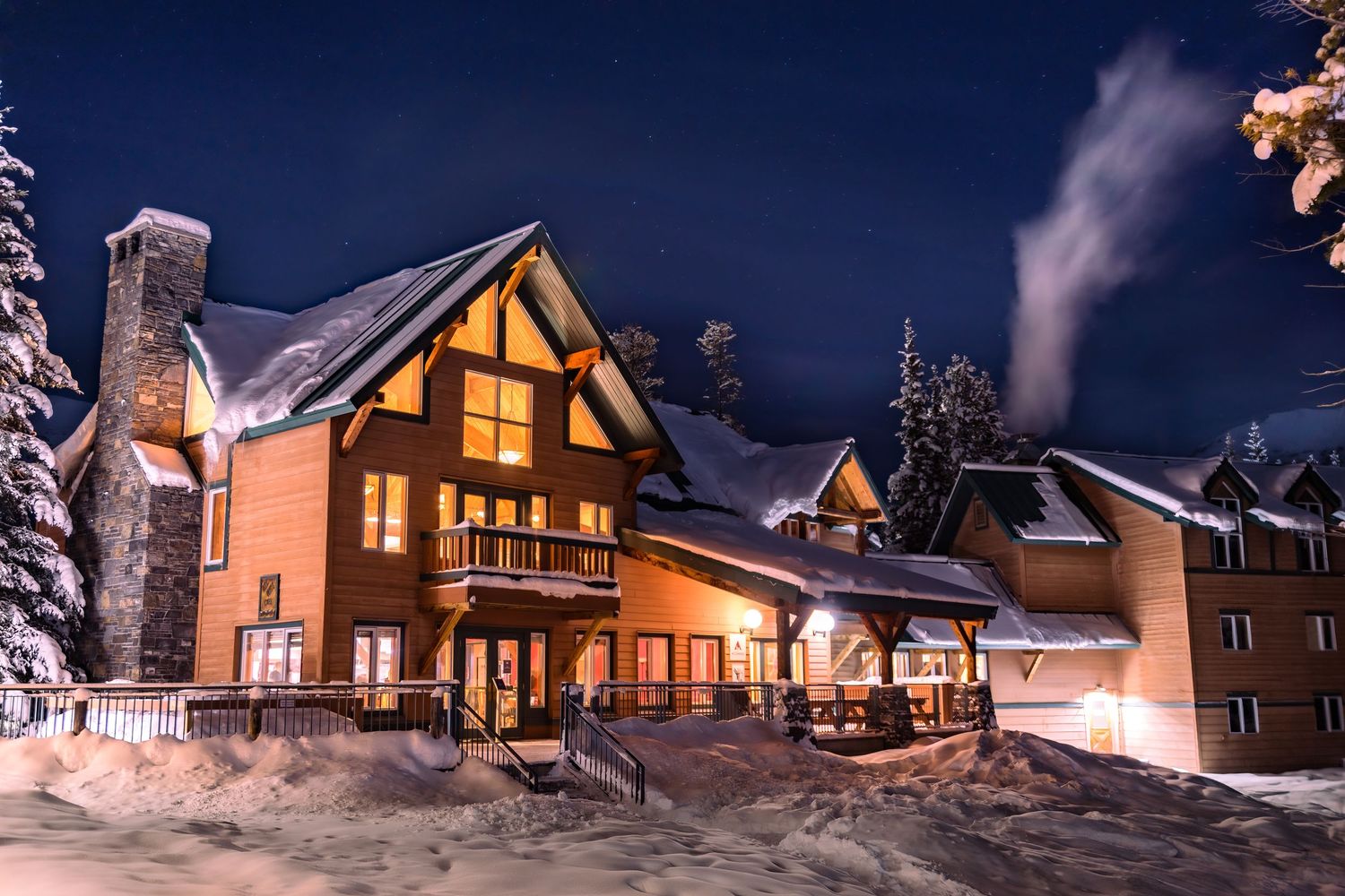 A row of illuminated wooden cabin lodges nestled in snowy forest during winter night creates a cozy mountain retreat scene.