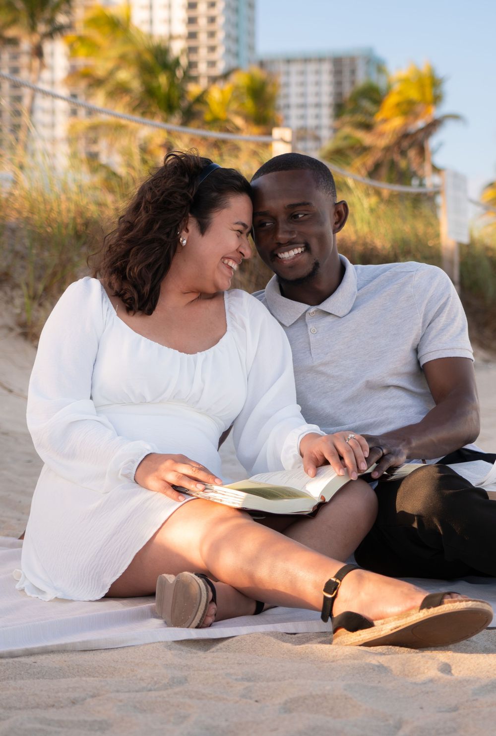 Happy interracial couple sitting on beach sand together reading a book at golden hour.