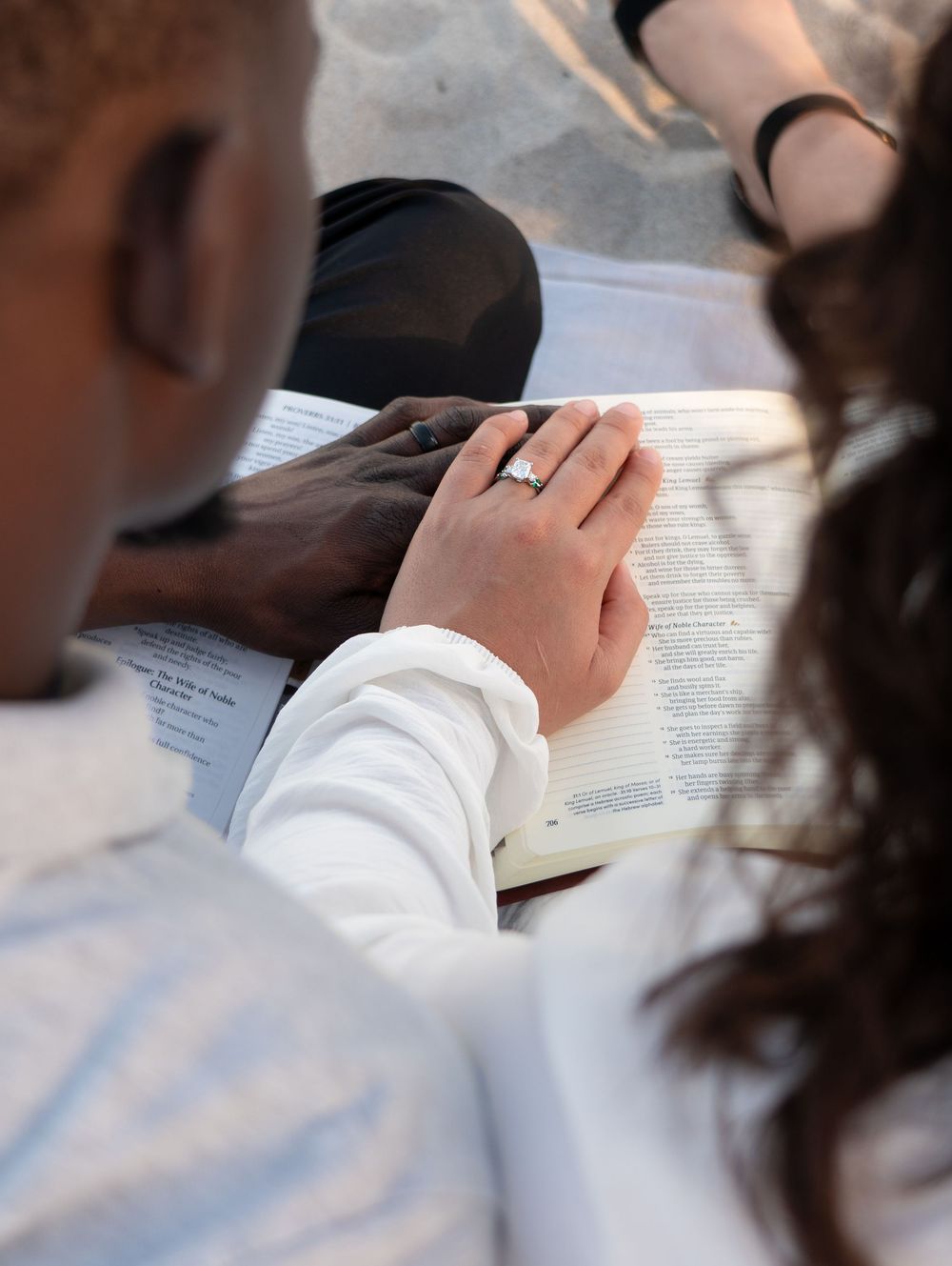 Close-up of a woman's hand with engagement ring resting on an open Bible shared by a couple.