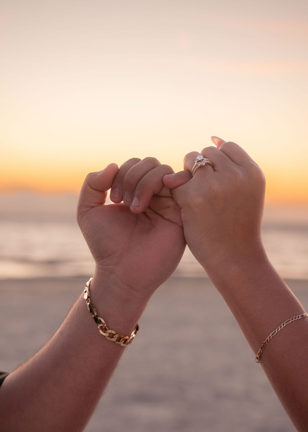 Two hands forming a heart shape at sunset on the beach, woman wearing an engagement ring.