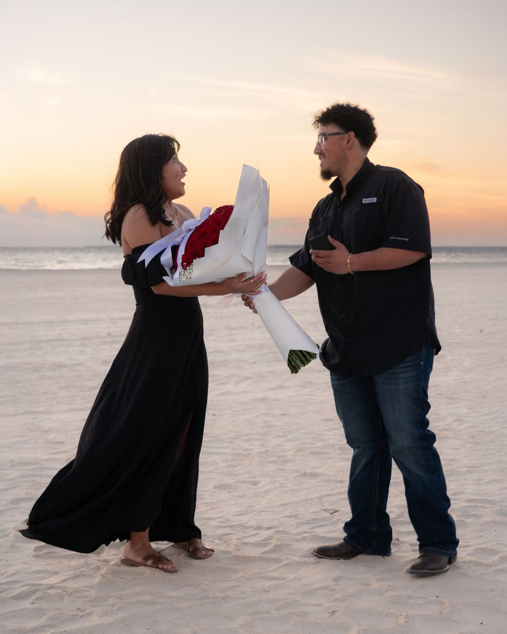 Man presenting a large bouquet of red roses to a woman in a black dress on the beach at sunset.