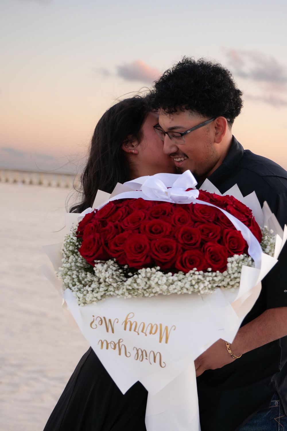 Couple kissing behind a large red rose bouquet with Marry Me sign at beach sunset.