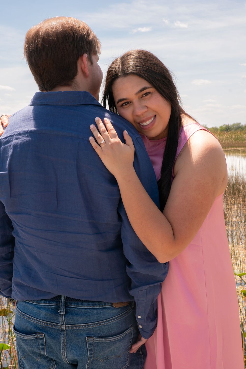 Smiling woman in pink dress hugging her partner outdoors near a scenic wetland backdrop.