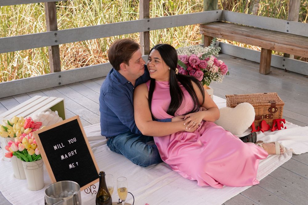 Couple sitting together at a romantic picnic proposal setup with Will You Marry Me sign.