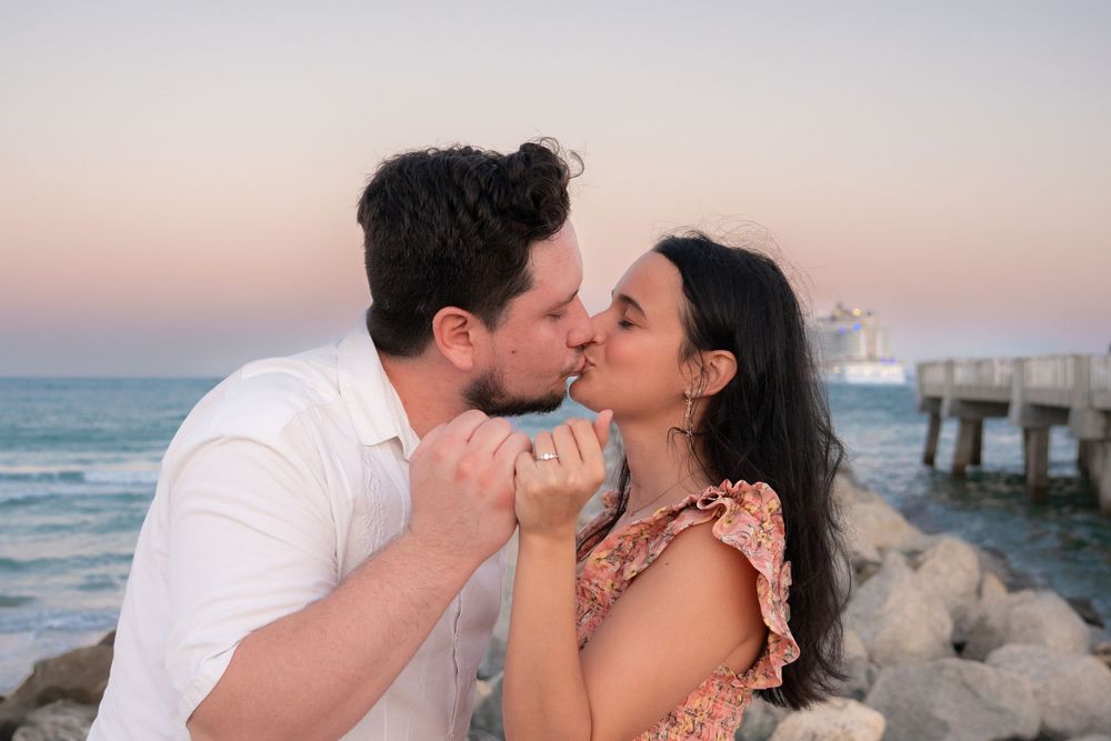 Couple sharing a tender kiss on rocky beach at sunset with pier in background.