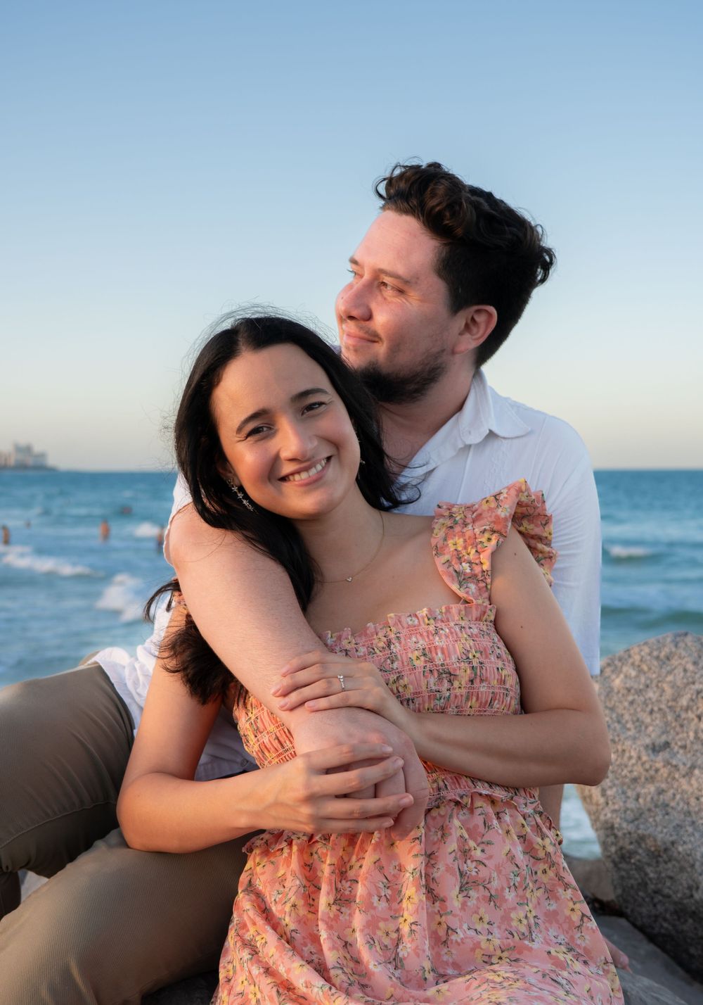 Happy couple embracing on beach rocks at dusk, woman in floral dress smiling warmly.