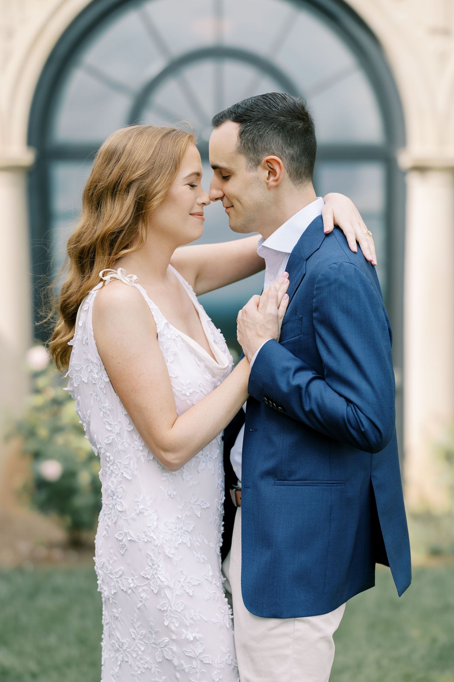 A romantic wedding couple shares an intimate moment in front of an arched doorway, wearing a lace gown and navy blazer at Pendry Natirar.