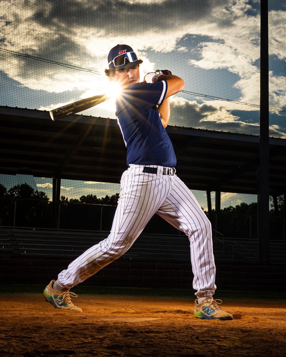 Baseball player in pinstripe uniform swings bat against dramatic sunset sky on baseball diamond with stadium lights.
