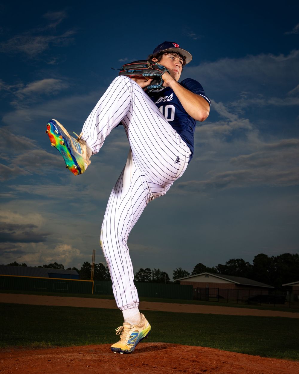 Baseball pitcher in pinstriped uniform performs dramatic high leg pitching motion against dramatic sky with clouds.