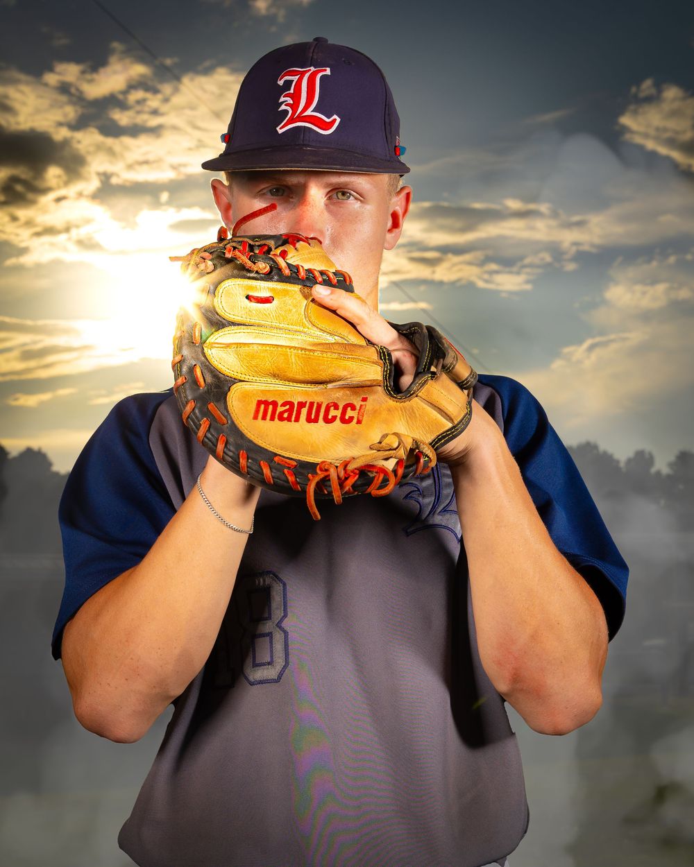 Baseball player in navy jersey and cap holds up Marucci glove against dramatic sunset sky background.