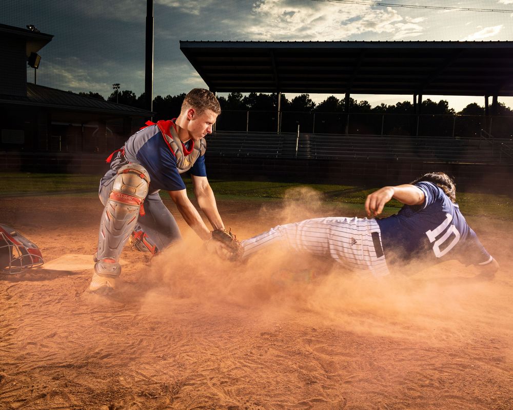 Two baseball players collide in dramatic play at home plate creating cloud of dirt on baseball diamond at sunset.