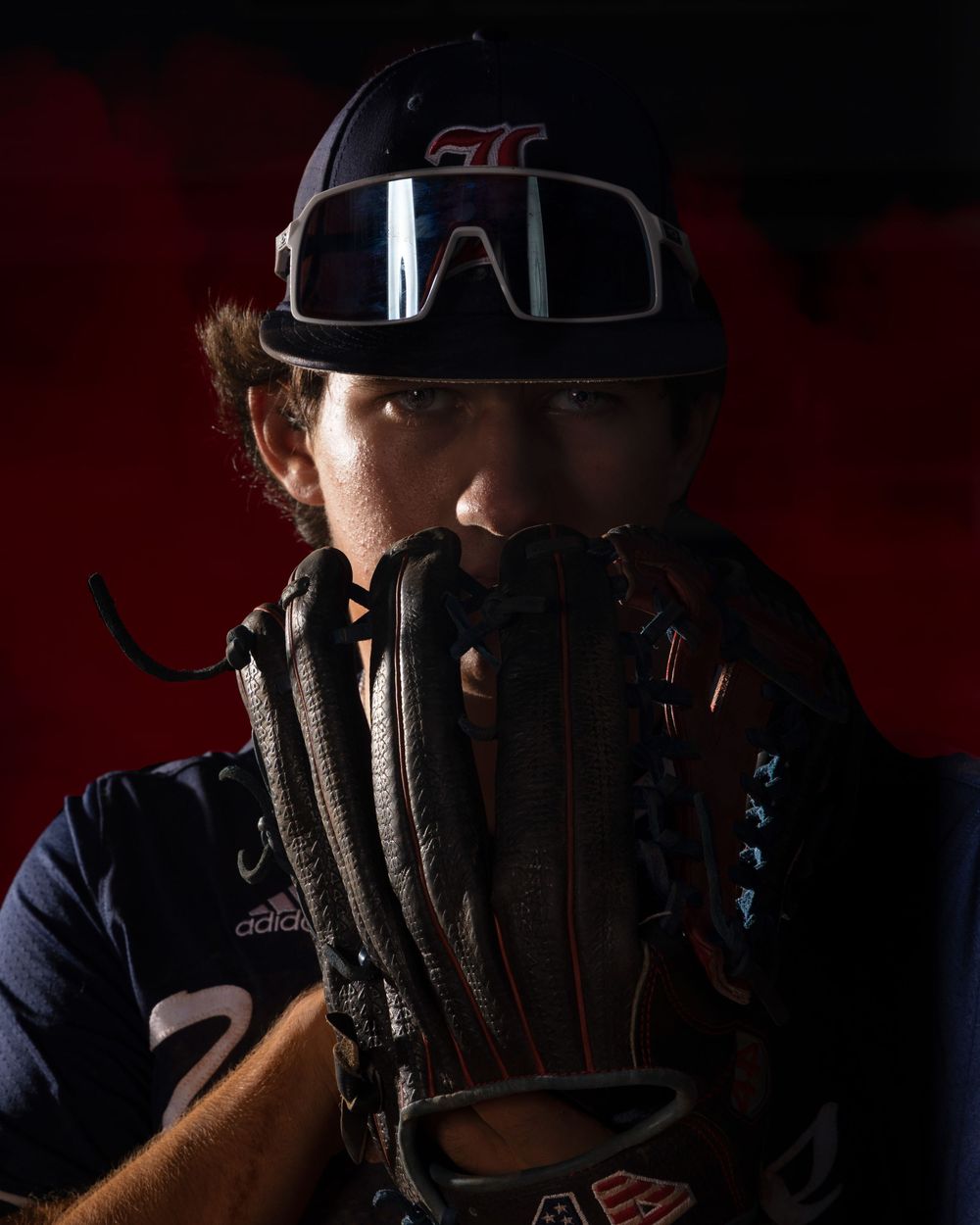 Baseball player in dark uniform peeks over their glove in dramatic lighting against red background.