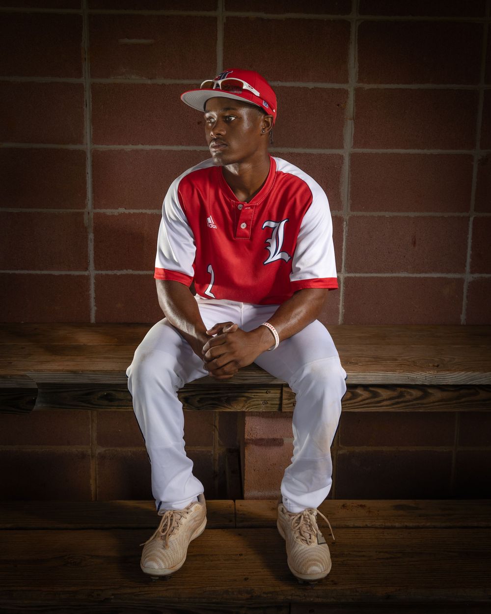 Baseball player in a red and white uniform sits in the dugout on wooden benches before a game.