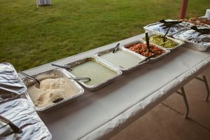 A buffet table set outdoors displays various food dishes in metal serving trays on a white tablecloth.